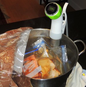 Nomiku sous vide immersion heater-circulator clipped to the side of a pot. Salmon and chicken underway. (I cover pot with plastic wrap and towels to minimize energy use.)